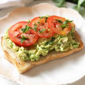 a slice of bread topped with mashed avocados on a white plate next to basil leaves.