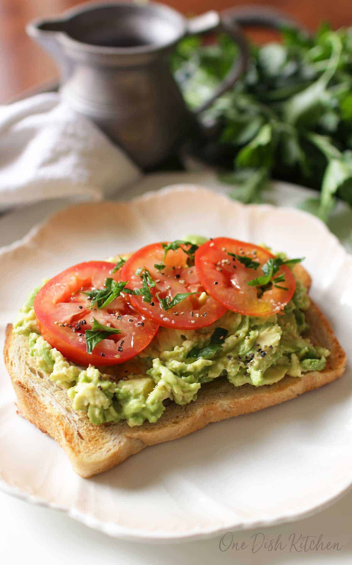 Avocado toast topped with three tomato slices, salt, pepper, and basil on a plate 