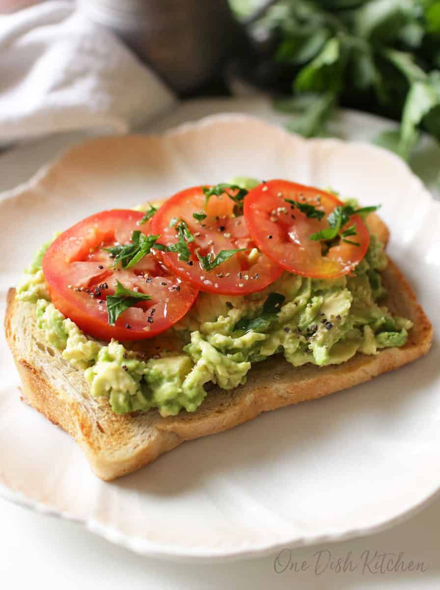 Avocado toast topped with three tomato slices, salt, pepper, and basil on a plate.