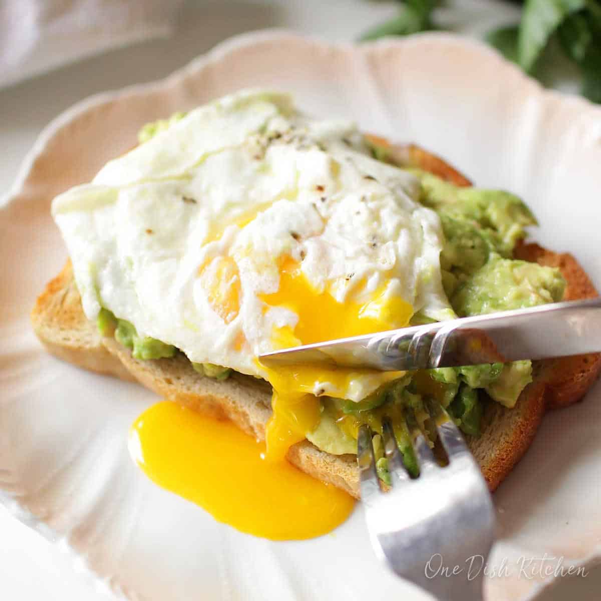 Fork and knife slicing into avocado toast with a runny fried egg.