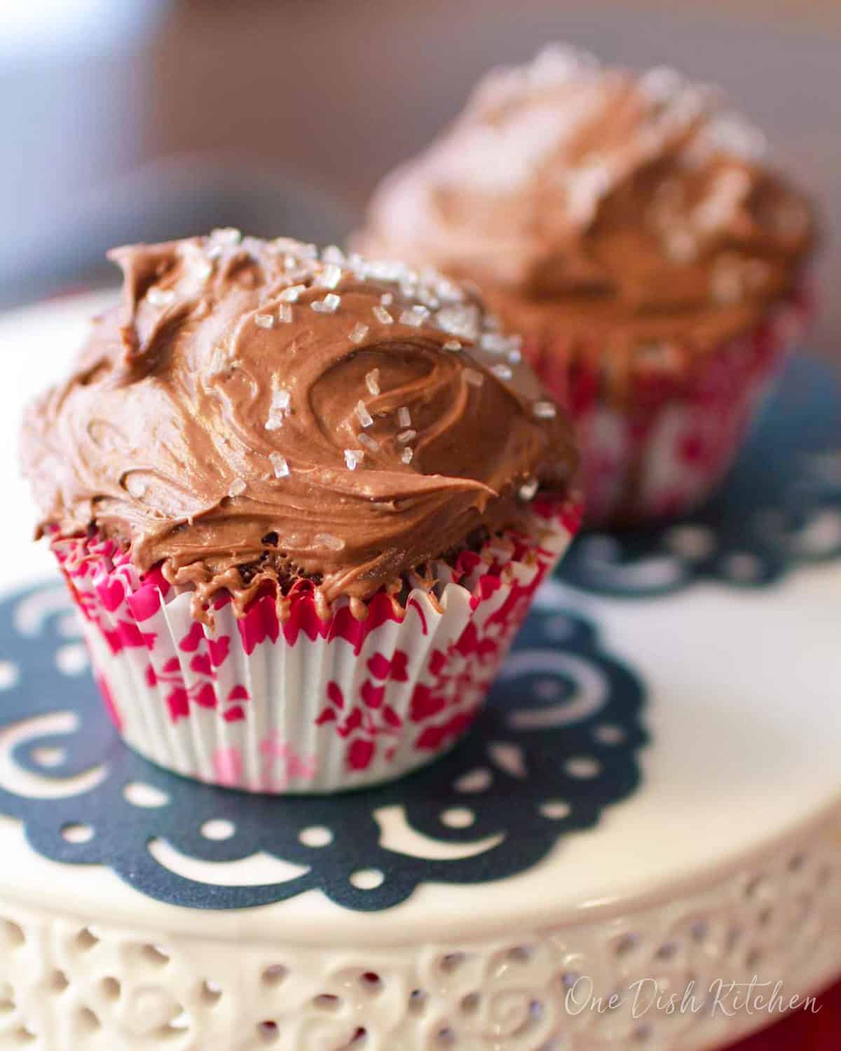 Two chocolate cupcakes with chocolate frosting and white crystal sprinkles on a cake stand.