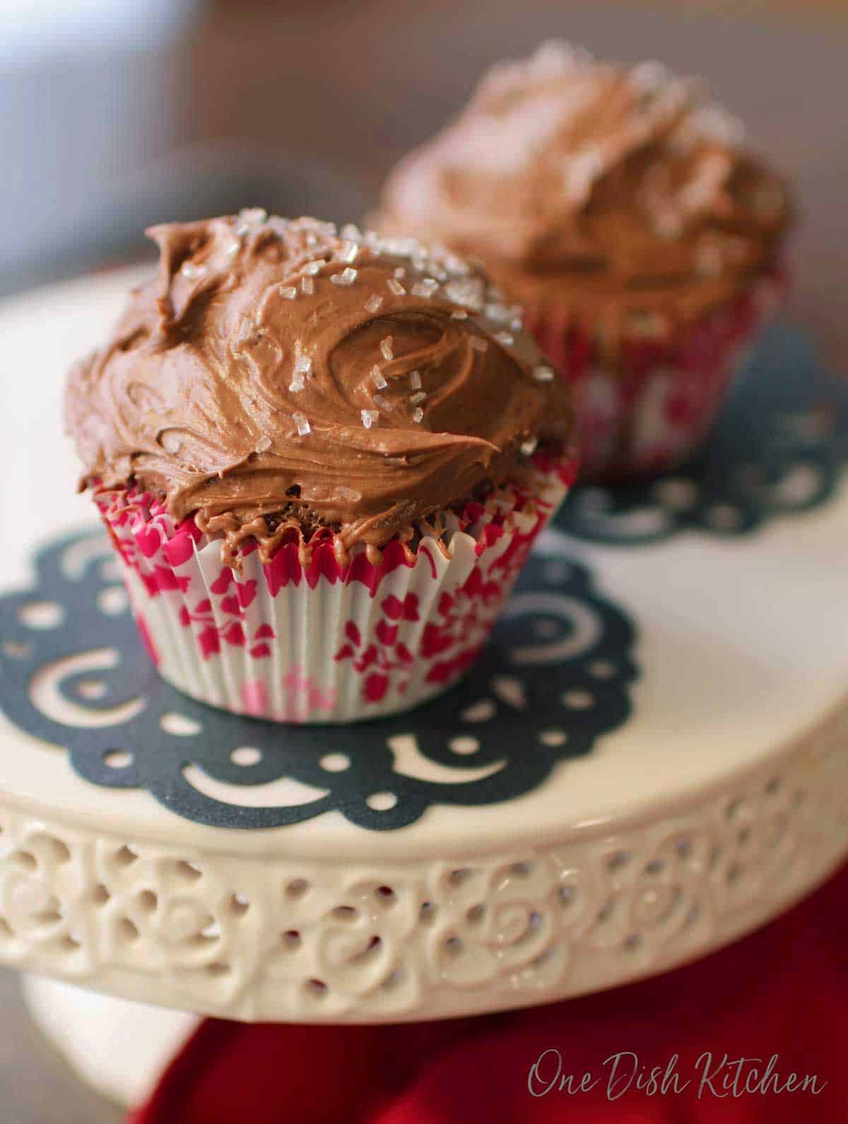 Two chocolate cupcakes with chocolate frosting and white crystal sprinkles on a cake stand.