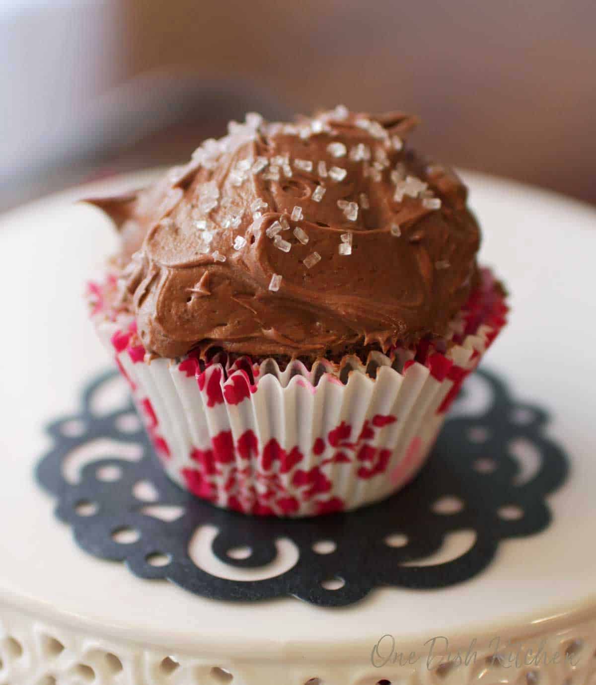 A chocolate cupcake topped with chocolate icing and white crystal sprinkles on a cake stand