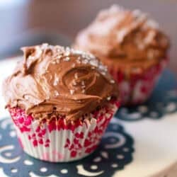 Two chocolate cupcakes with chocolate frosting and white crystal sprinkles on a cake stand.