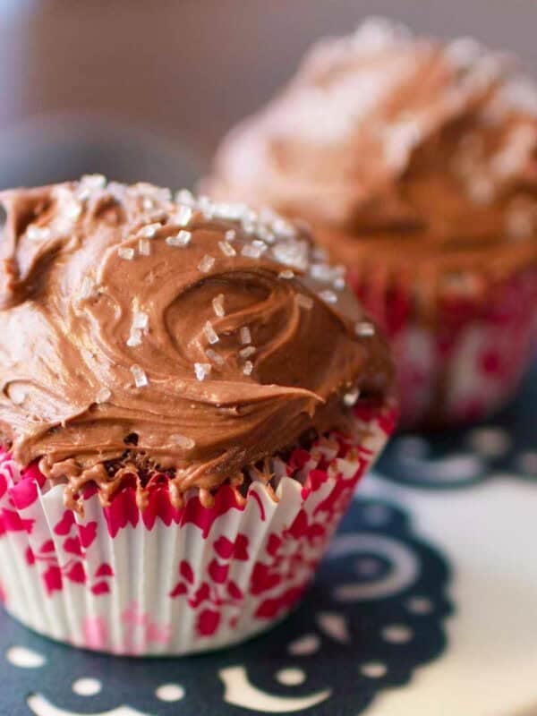 Two chocolate cupcakes with chocolate frosting and white crystal sprinkles on a cake stand.