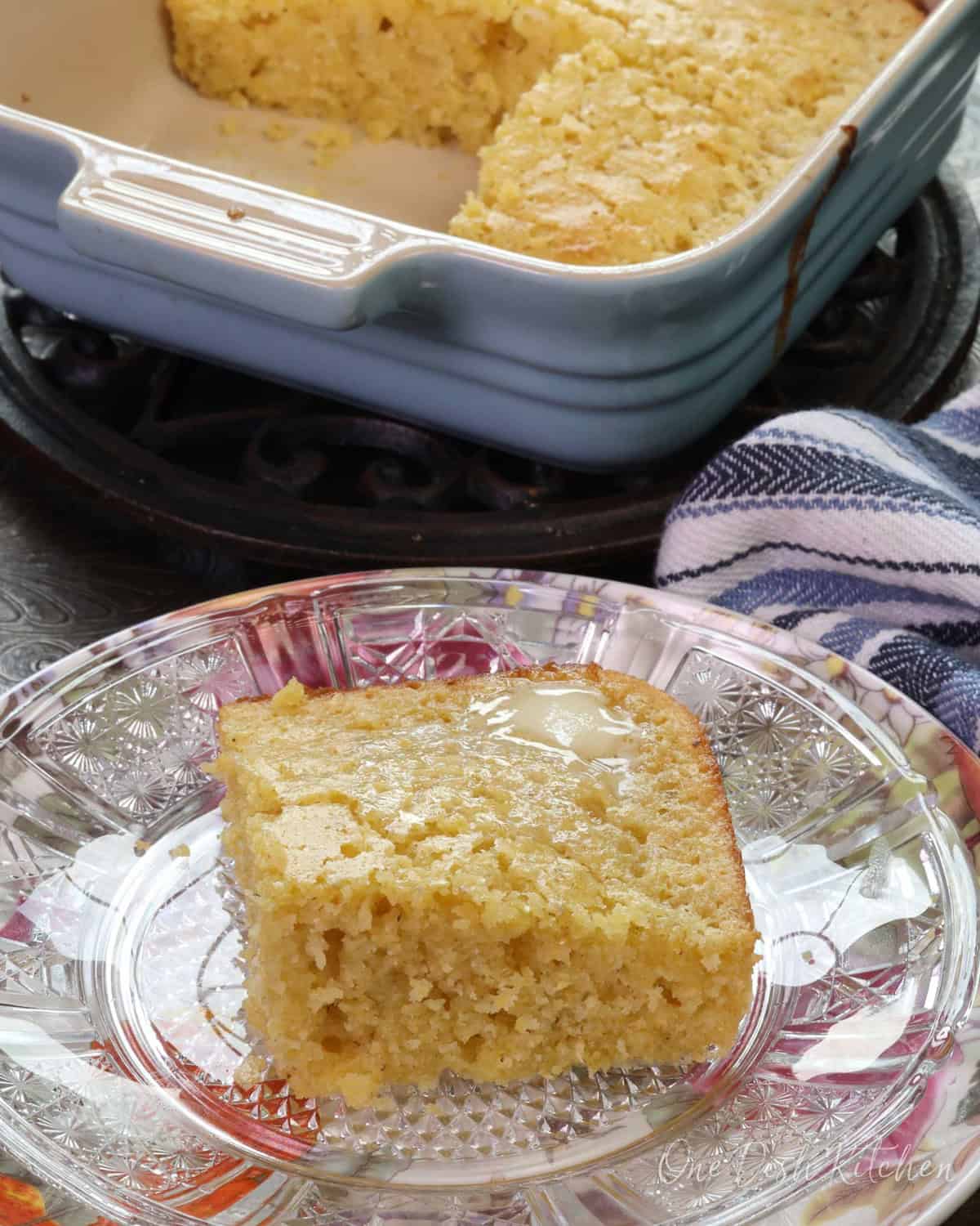 a square piece of cornbread on a floral plate next to a pan filled with three squares of cornbread.