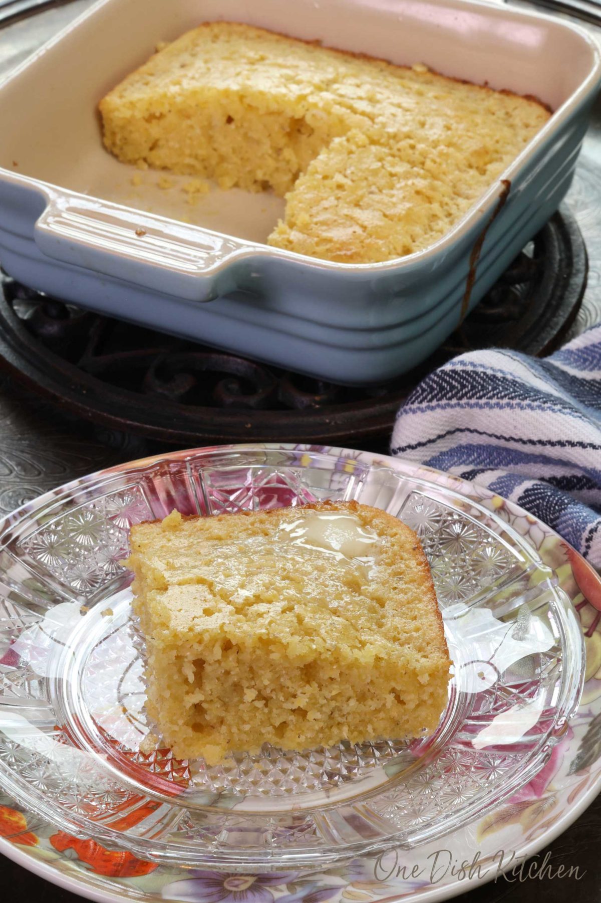 a square piece of cornbread on a floral plate next to a pan filled with three squares of cornbread.