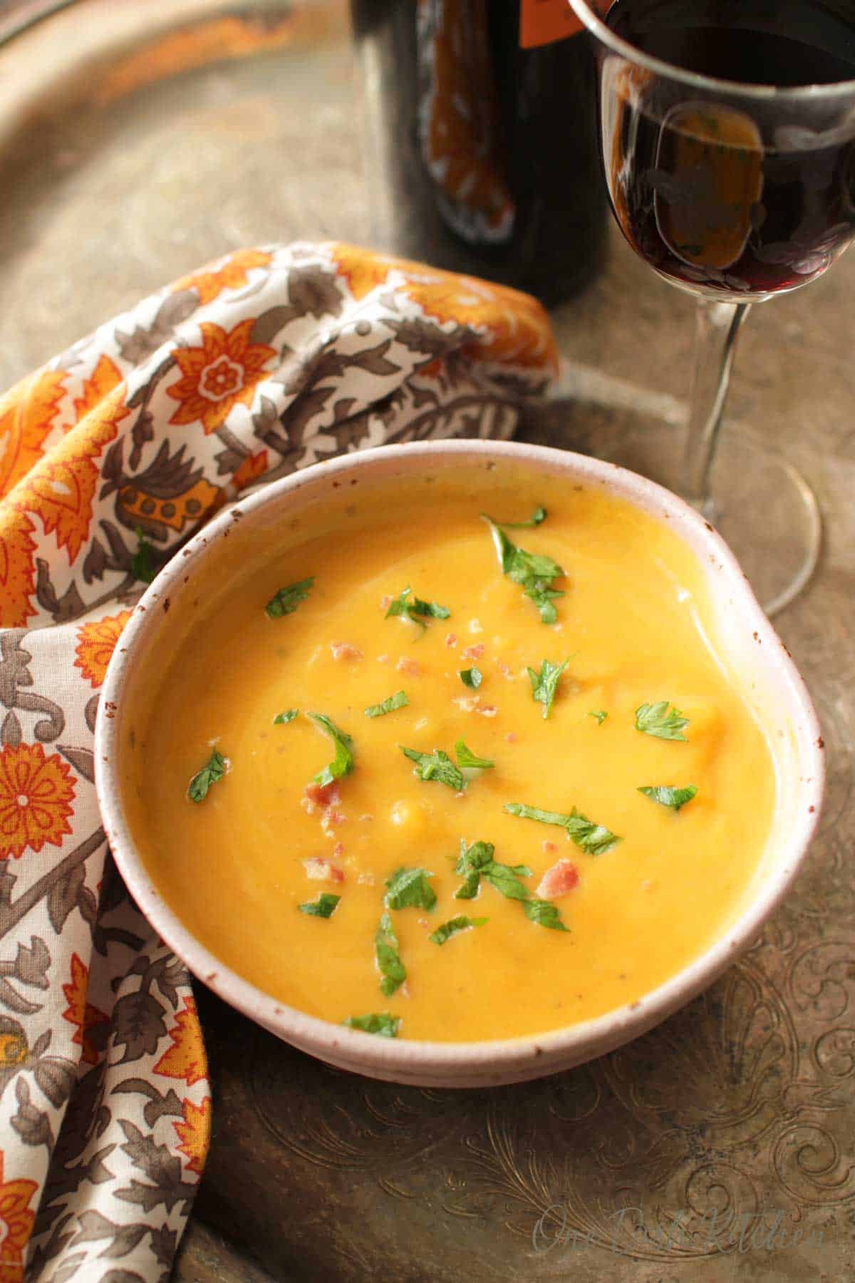 Butternut Squash Soup in a bowl next to a glass of red wine and an orange and grey cloth napkin all on a metal tray.
