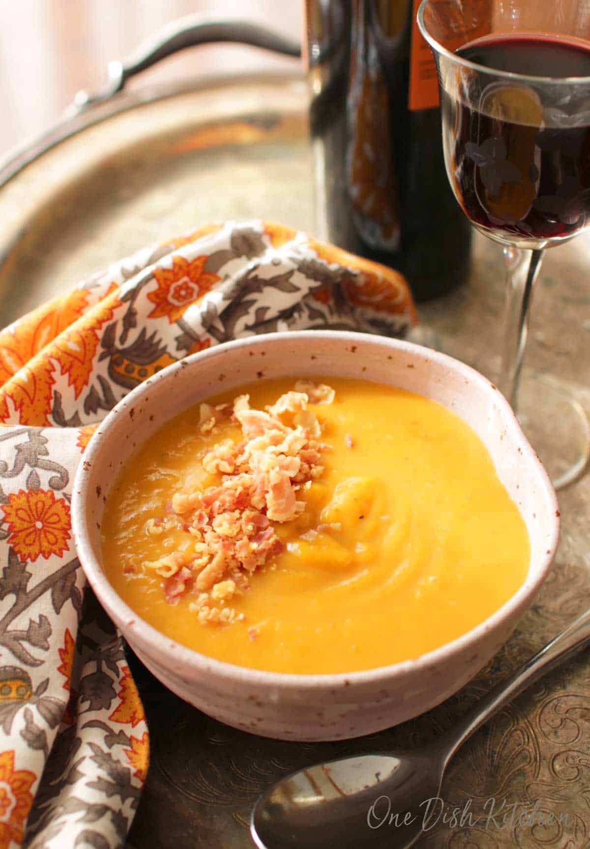 A bowl of butternut squash soup topped with homemade croutons next to a glass and bottle of red wine and a orange and grey cloth napkin all on a metal tray.