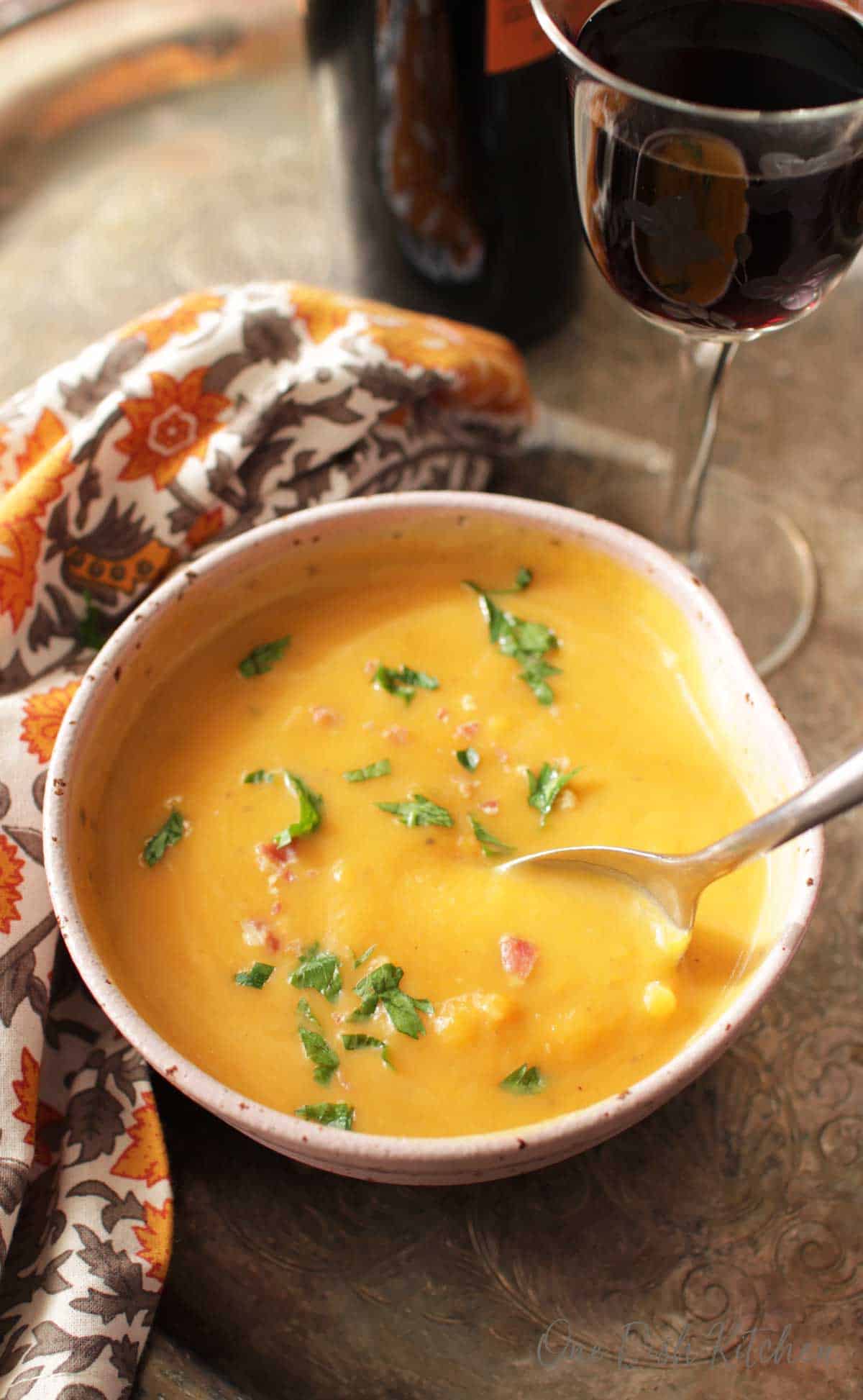 An overhead view of a spoon diving into a bowl of Butternut Squash Soup next to a glass of red wine and an orange and grey cloth napkin all on a metal tray.
