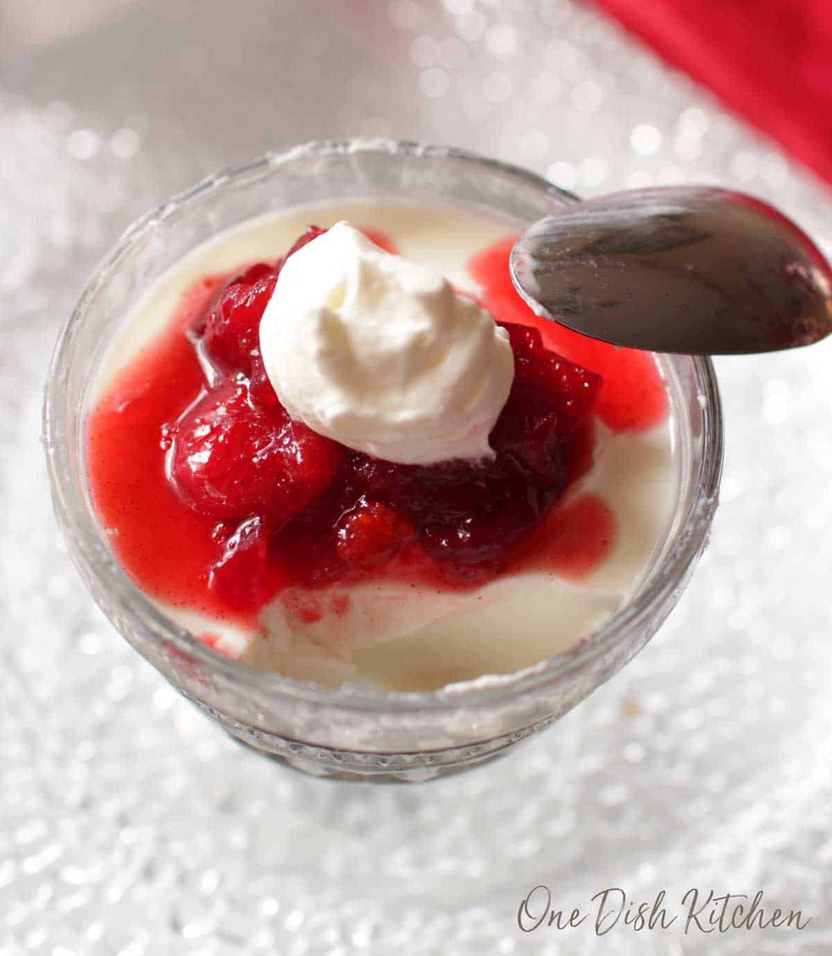 An overhead view of panna cotta topped with cranberry jam and whipped cream in a dessert glass with a spoon resting on the side of the glass