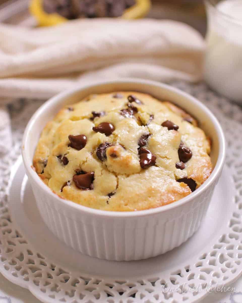 A chocolate chip muffin in a ramekin next to a yellow dish filled with chocolate chips and a small glass of milk.
