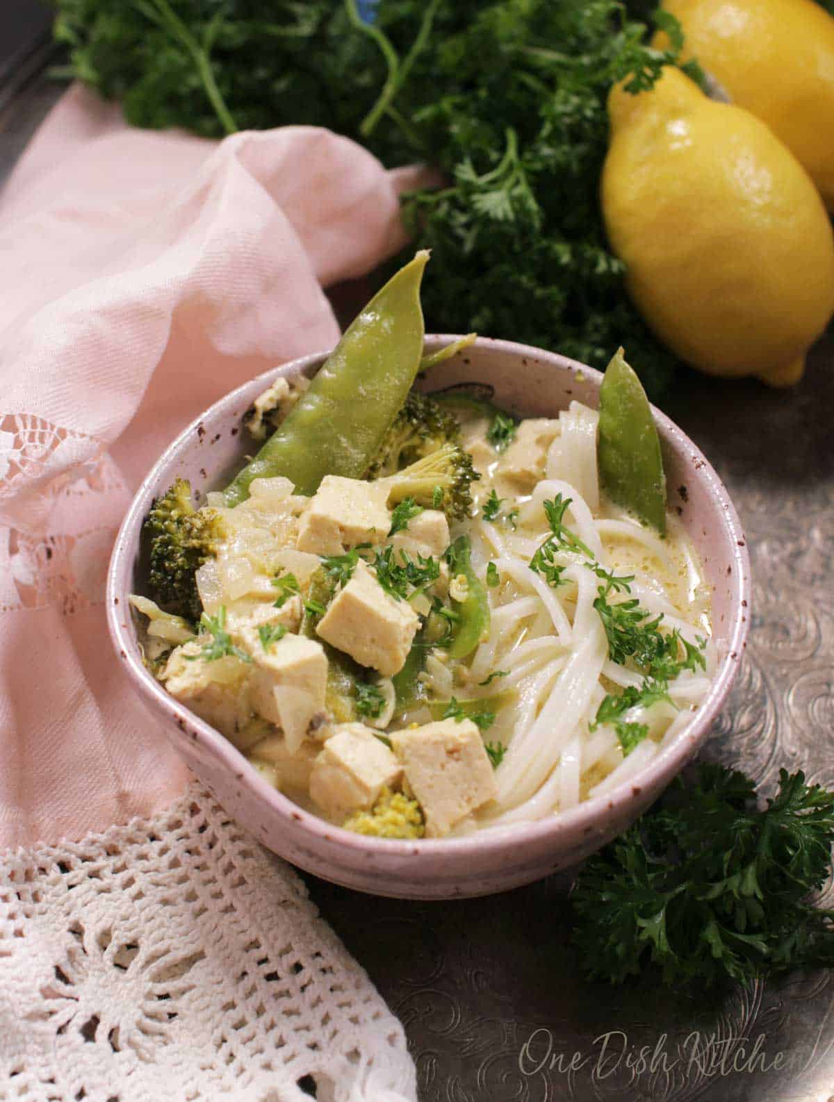 A bowl of green curry with tofu and noodles on a metal tray with a pink cloth napkin, parsley, and two lemons.