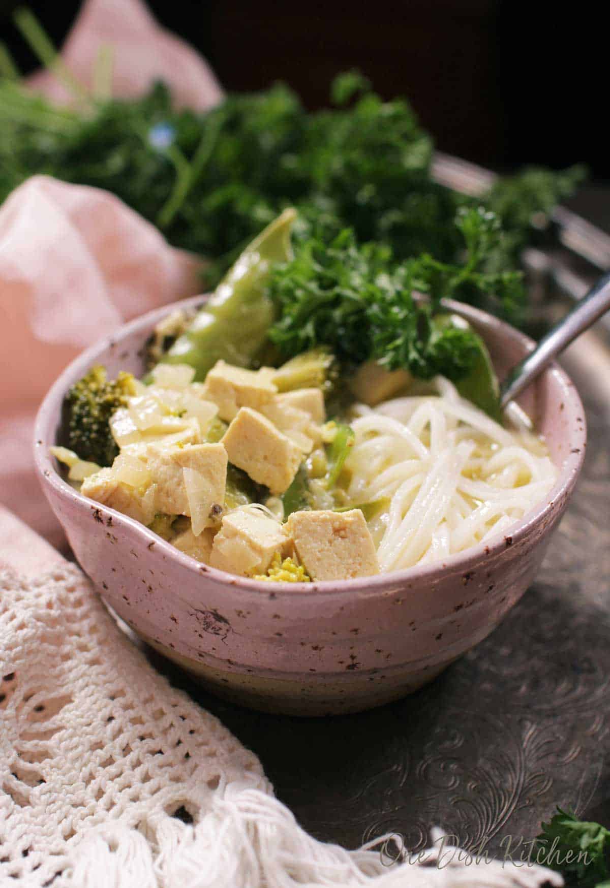 A closeup of a bowl of green curry with tofu and noodles on a metal tray  with parsley.