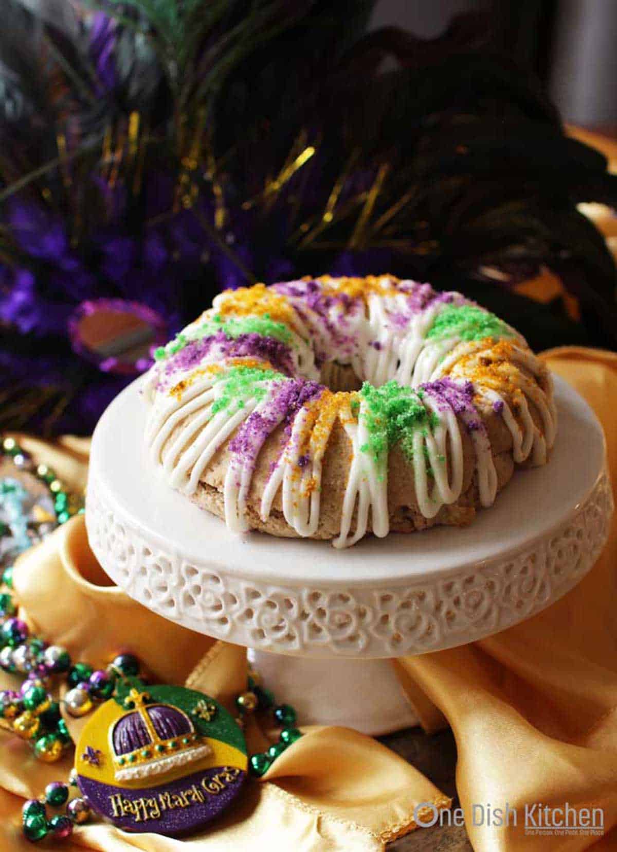 A mini king cake topped with white drizzled icing and green, orange, and purple sugar on a cake stand surrounded by a gold cloth and mardi gras beads.