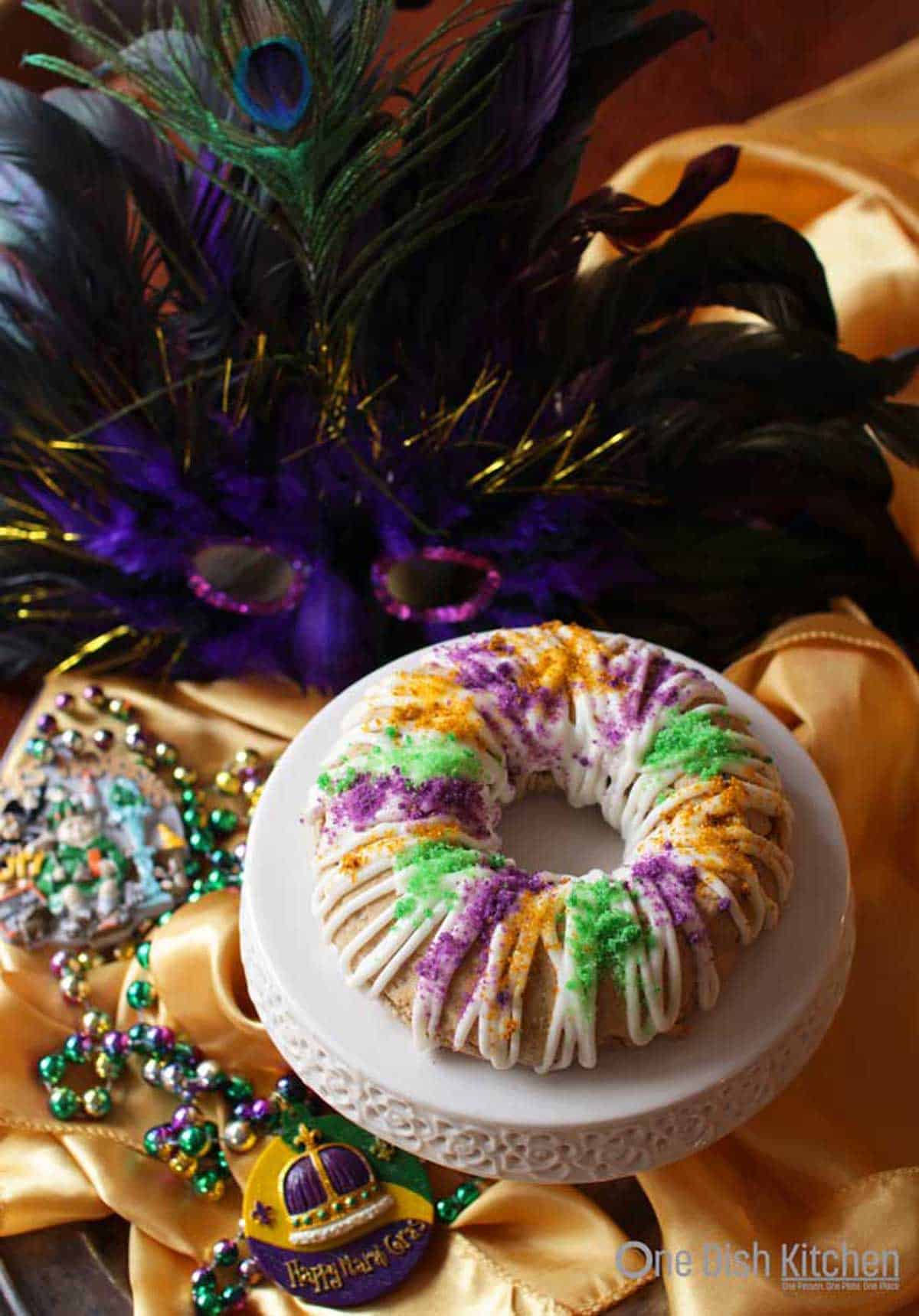 An overhead view of a mini king cake topped with white drizzled icing and green, orange, and purple sugar on a cake stand surrounded by a gold cloth, mardi gras beads, and a purple and gold feather mask 