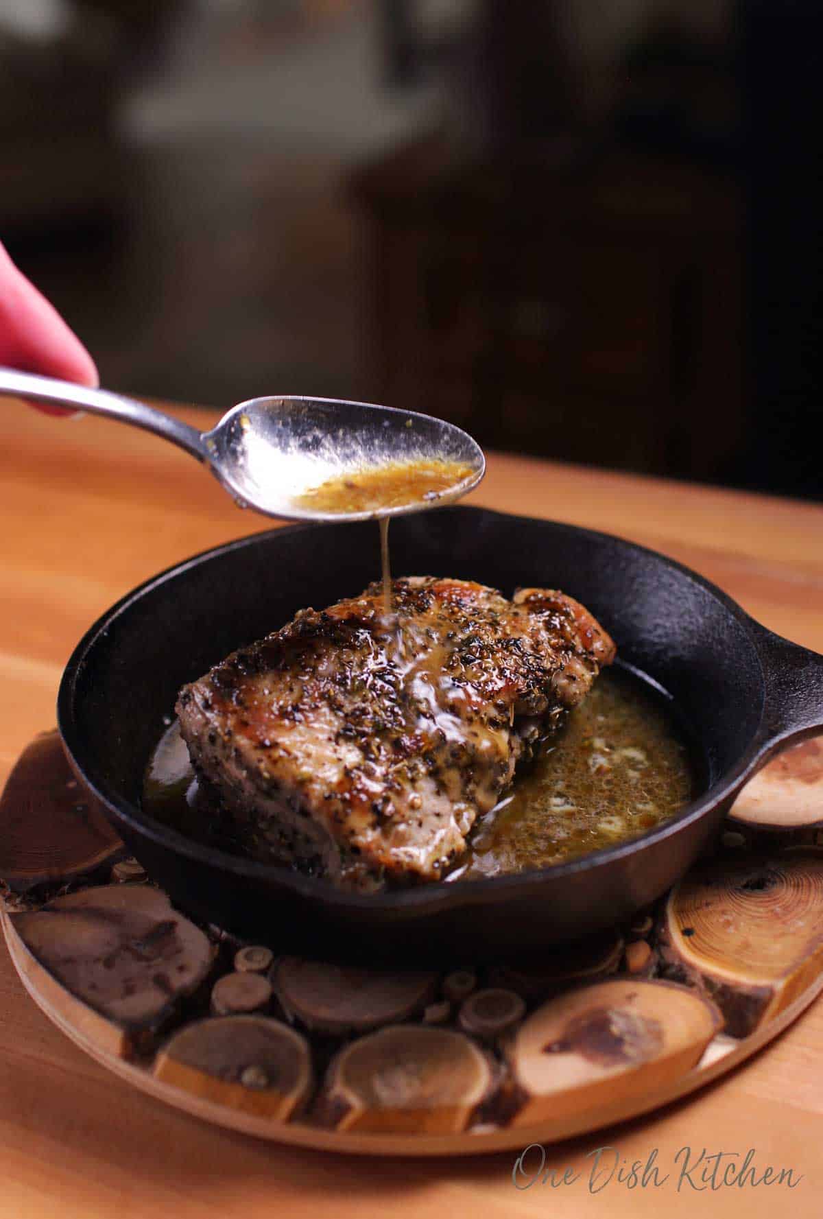 A spoon pouring gravy onto a pork tenderloin in a cast iron skillet on a wooden trivet.