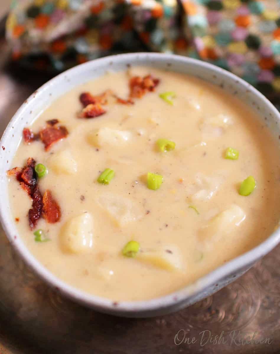 A closeup of a bowl of creamy potato soup topped with chopped bacon next to a spoon all on a metal tray.
