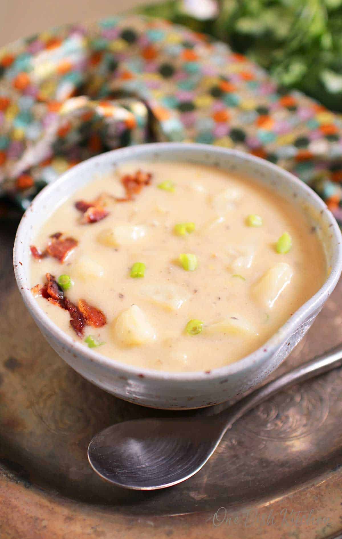 A closeup of a bowl of creamy potato soup topped with chopped bacon next to a spoon all on a metal tray.
