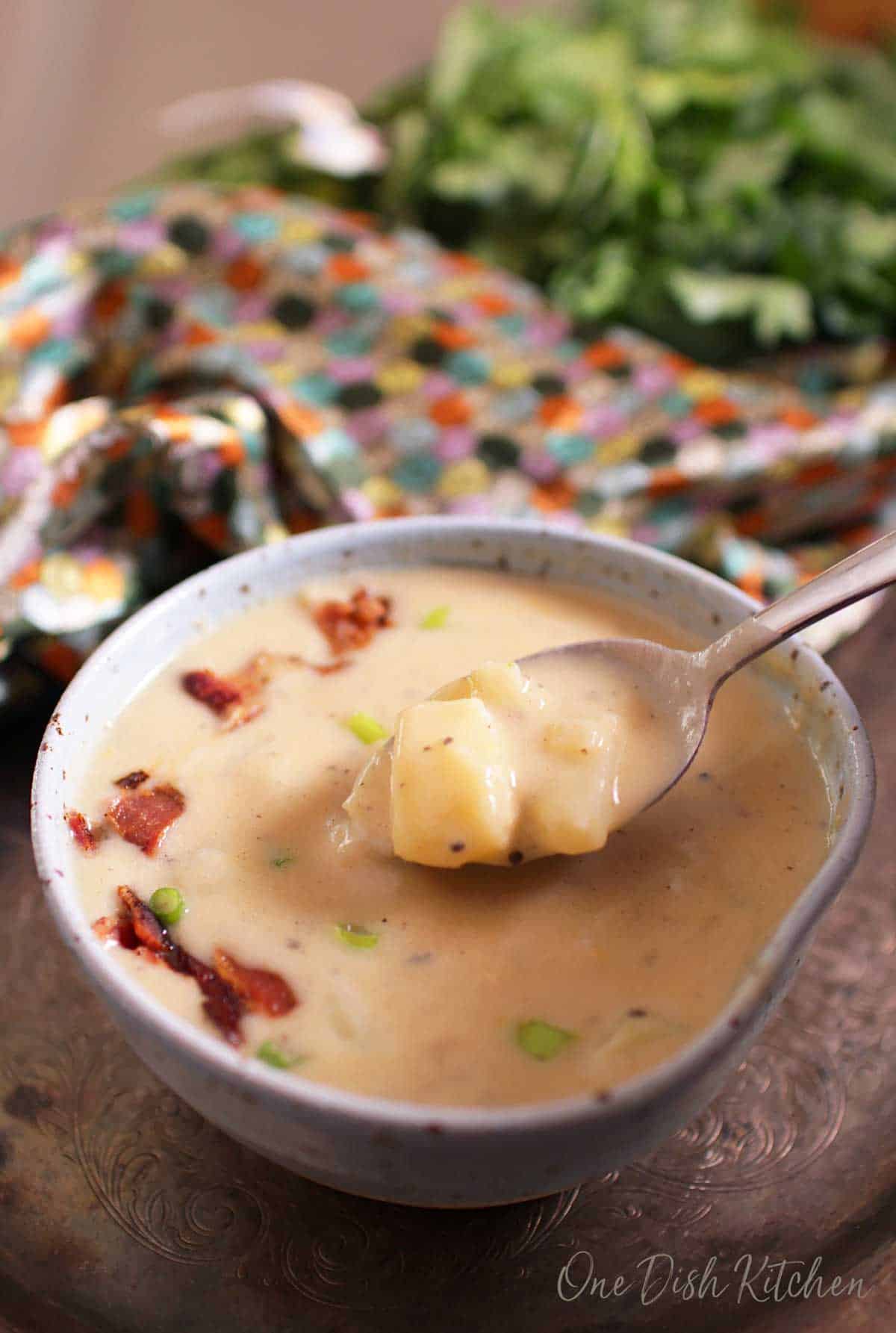 A spoonful of potato soup topped with chopped bacon from a bowl on a metal tray next to a multi-colored cloth napkin.