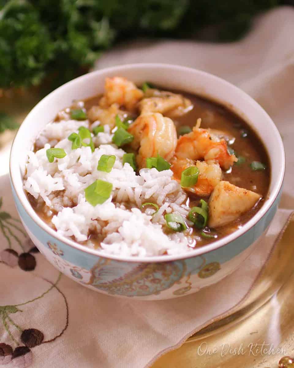 A bowl of gumbo with shrimp and white rice on a tray with parsley in the background.