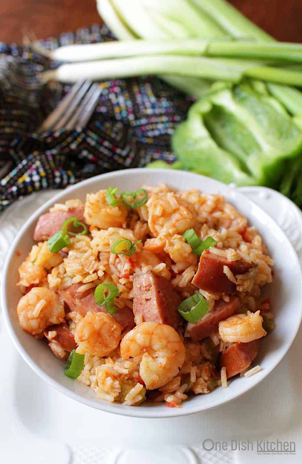 Shrimp and sausage jambalaya in a bowl with onions, celery, and green bell peppers in the background.