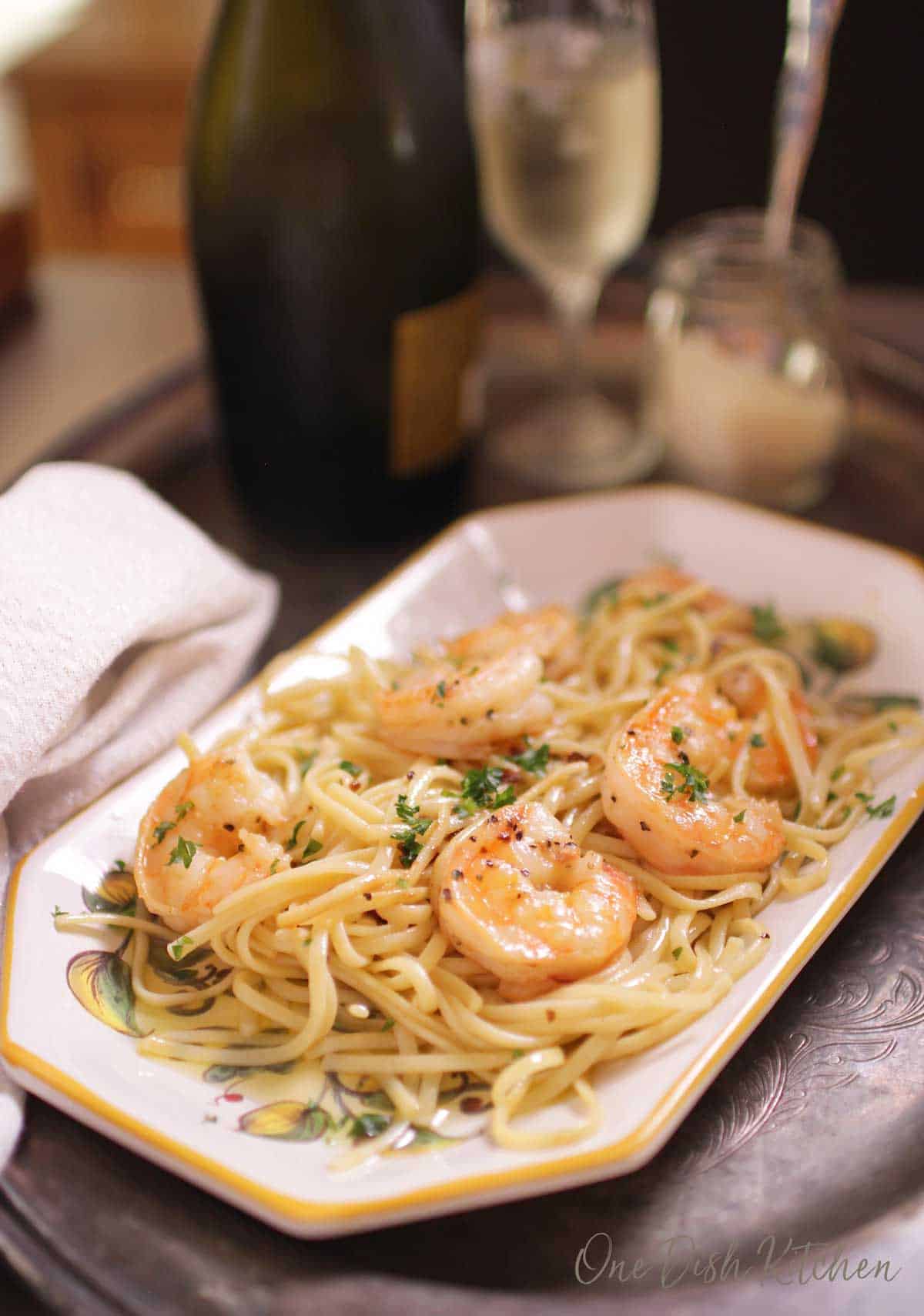 A plate of spaghetti with shrimp on a tray next to a small jar of parmesan cheese and a glass of champagne.