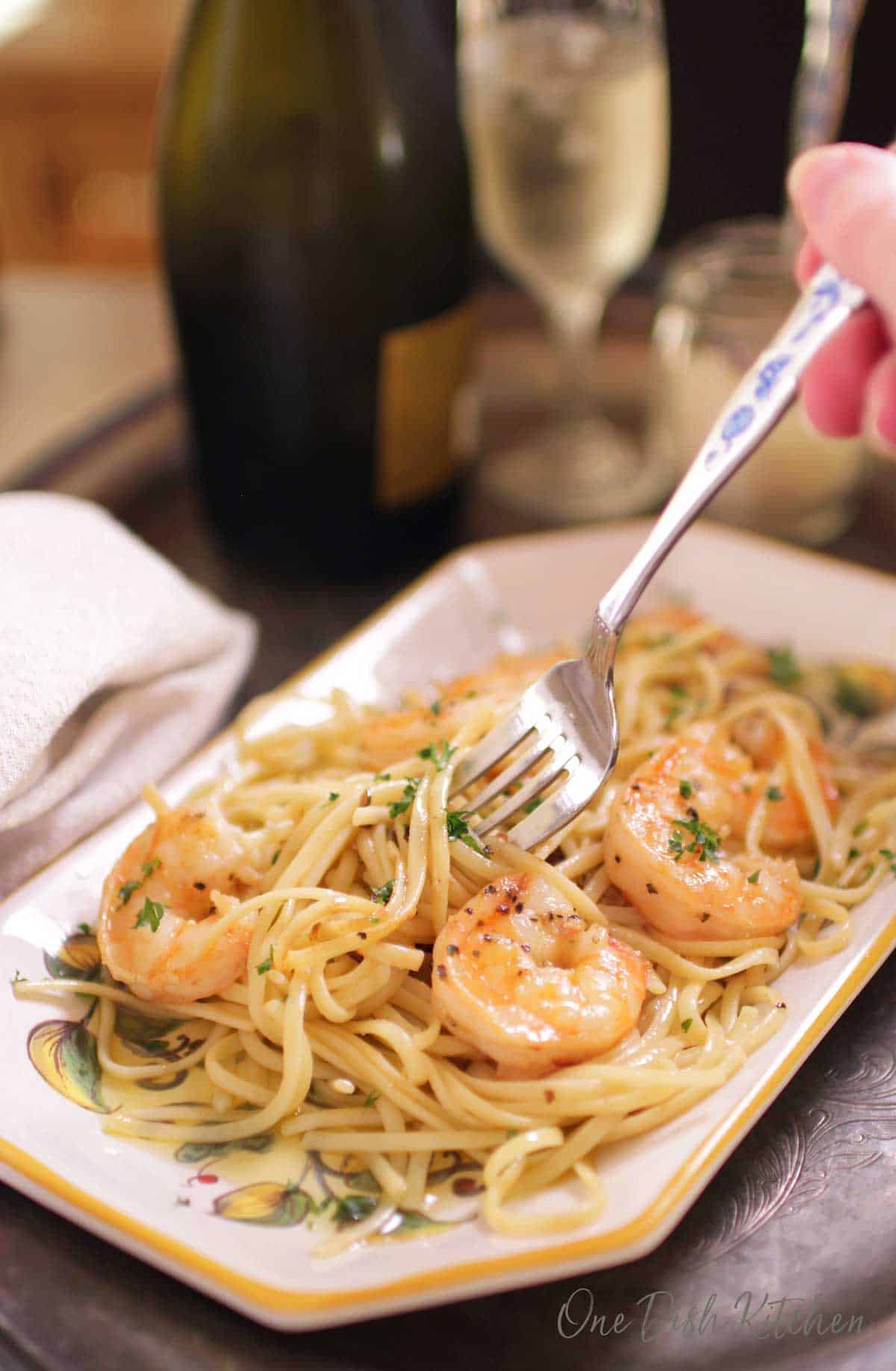 A fork in shrimp scampi on a plate next to a glass of champagne all on a metal tray