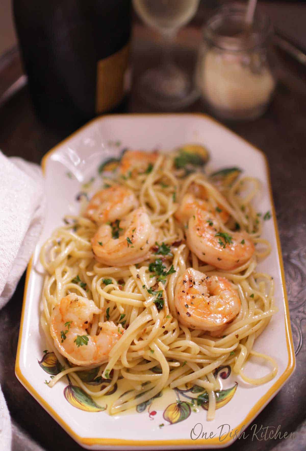An overhead view of a plate of spaghetti with shrimp on a tray next to a small jar of parmesan cheese and a glass of champagne.
