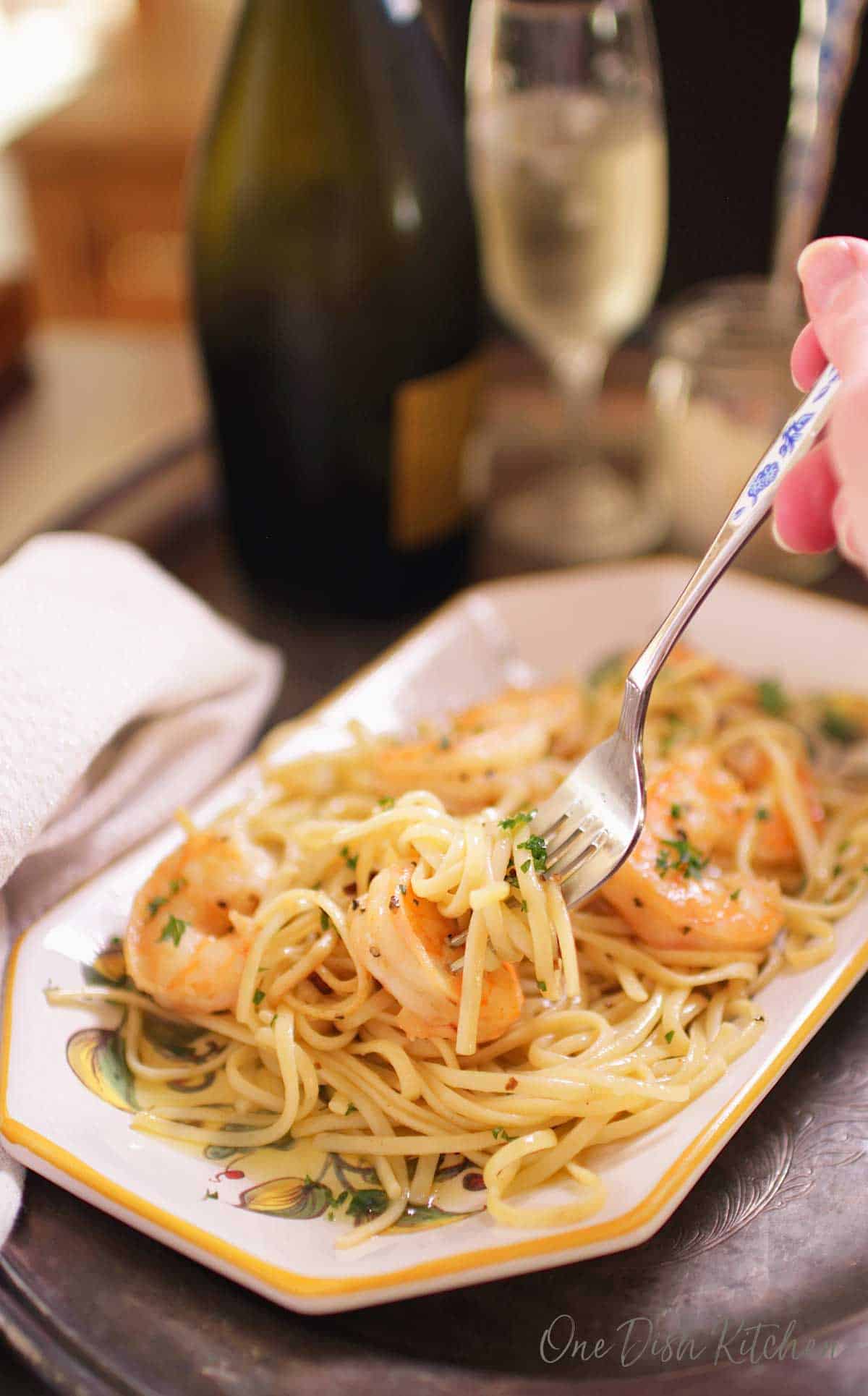 A fork in shrimp scampi on a plate next to a glass of champagne all on a metal tray