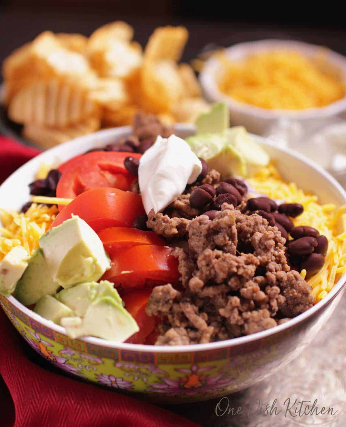 A closeup of a bowl of taco salad filled with ground beef, tomatoes, avocados and cheese topped with sour cream 