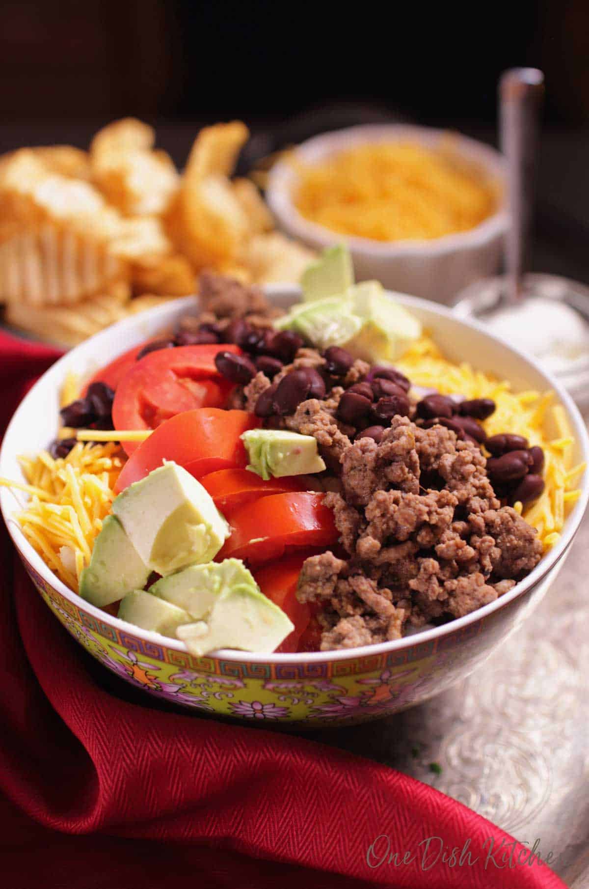 A bowl of taco salad filled with ground beef, tomatoes, avocados and cheese next to a bowl of chips.