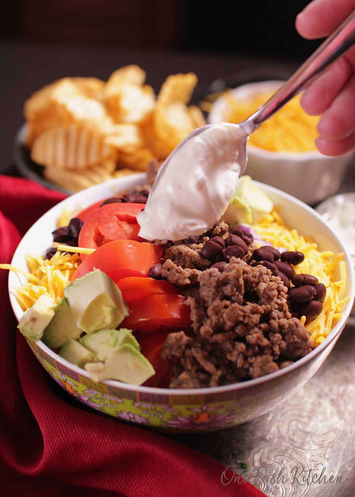 Placing a dollop of sour cream on top of a bowl of taco salad filled with ground beef, tomatoes, avocados and cheese next to a bowl of chips.