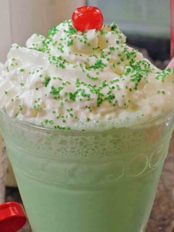 a green colored shamrock shake with whipped topping and cheery on top in a decorative glass.