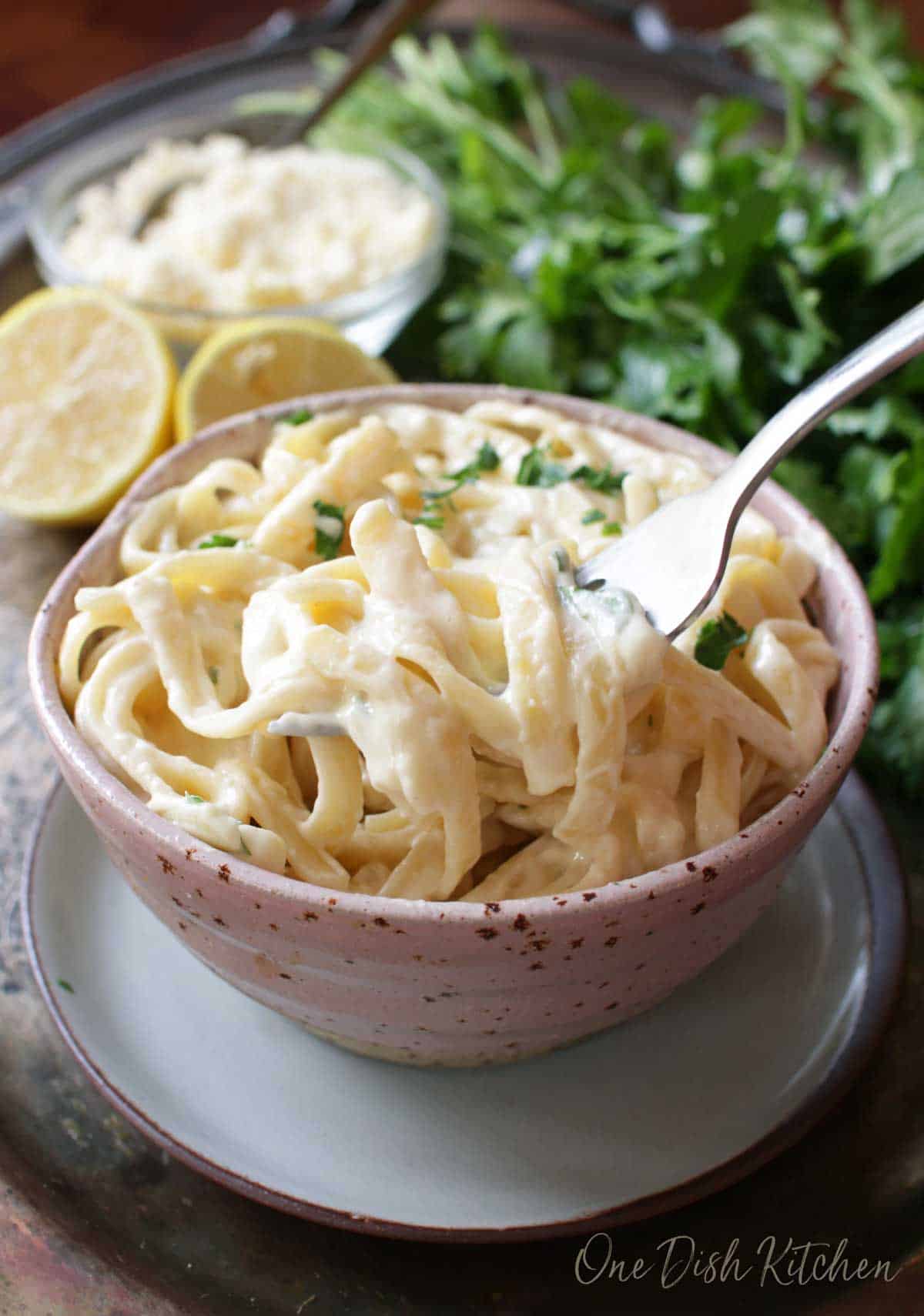 A forkful of fettuccine alfredo from the bowl next to a dish of grated parmesan cheese and lemon halves all on a metal tray