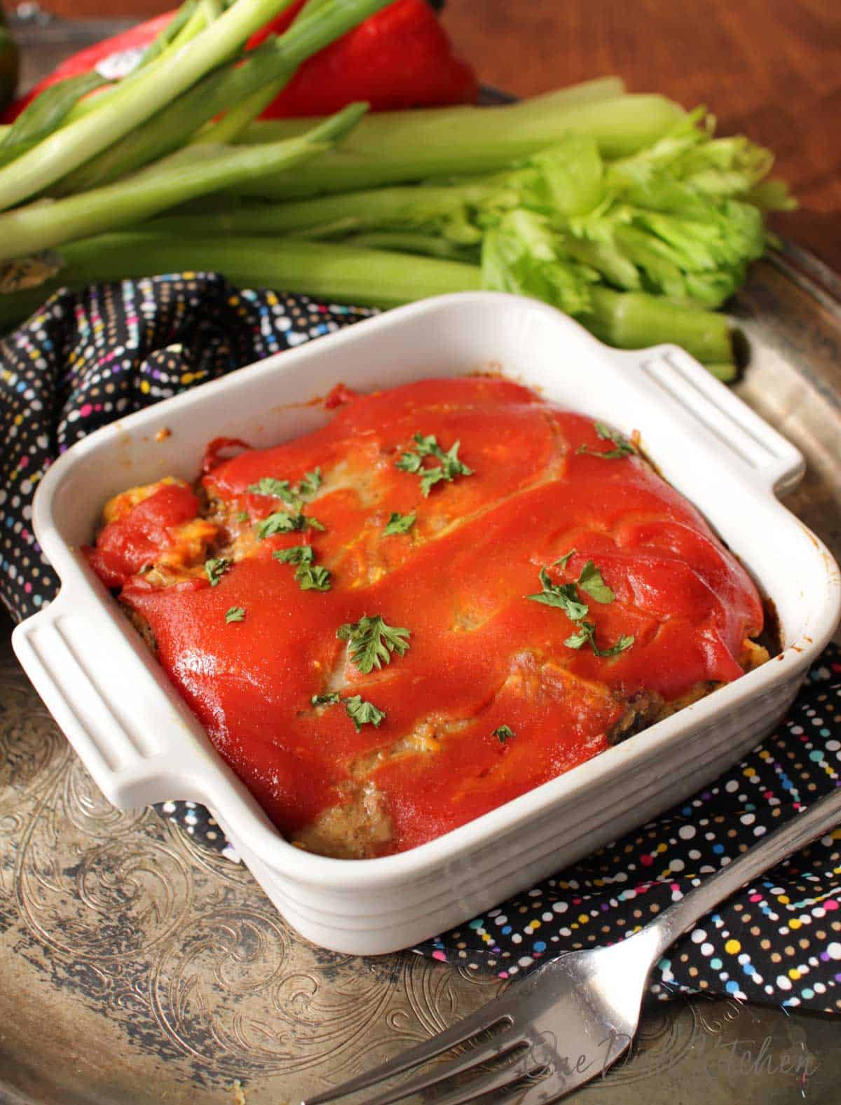 A single serving bacon cheeseburger meatloaf in a small baking dish next to a fork and bunch of celery all on a metal tray 