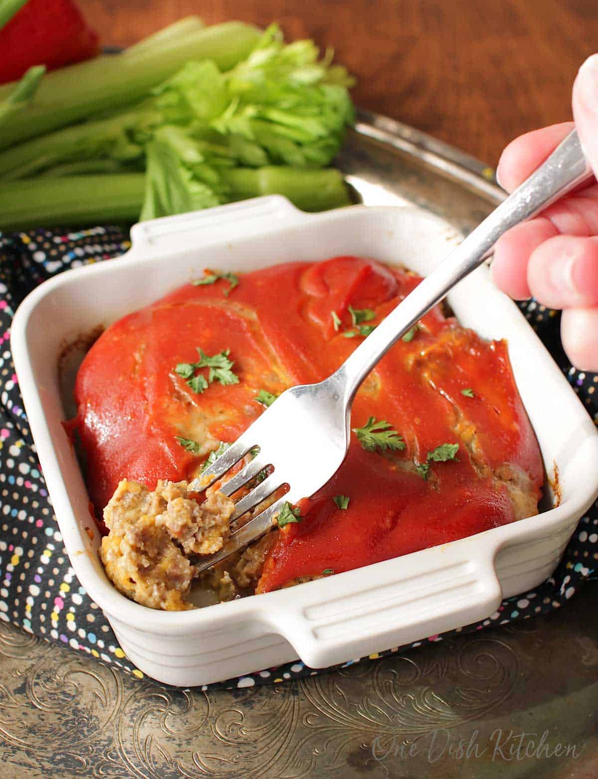 A forkful of bacon cheeseburger meatloaf from a small baking dish next to a bunch of celery all on a metal tray