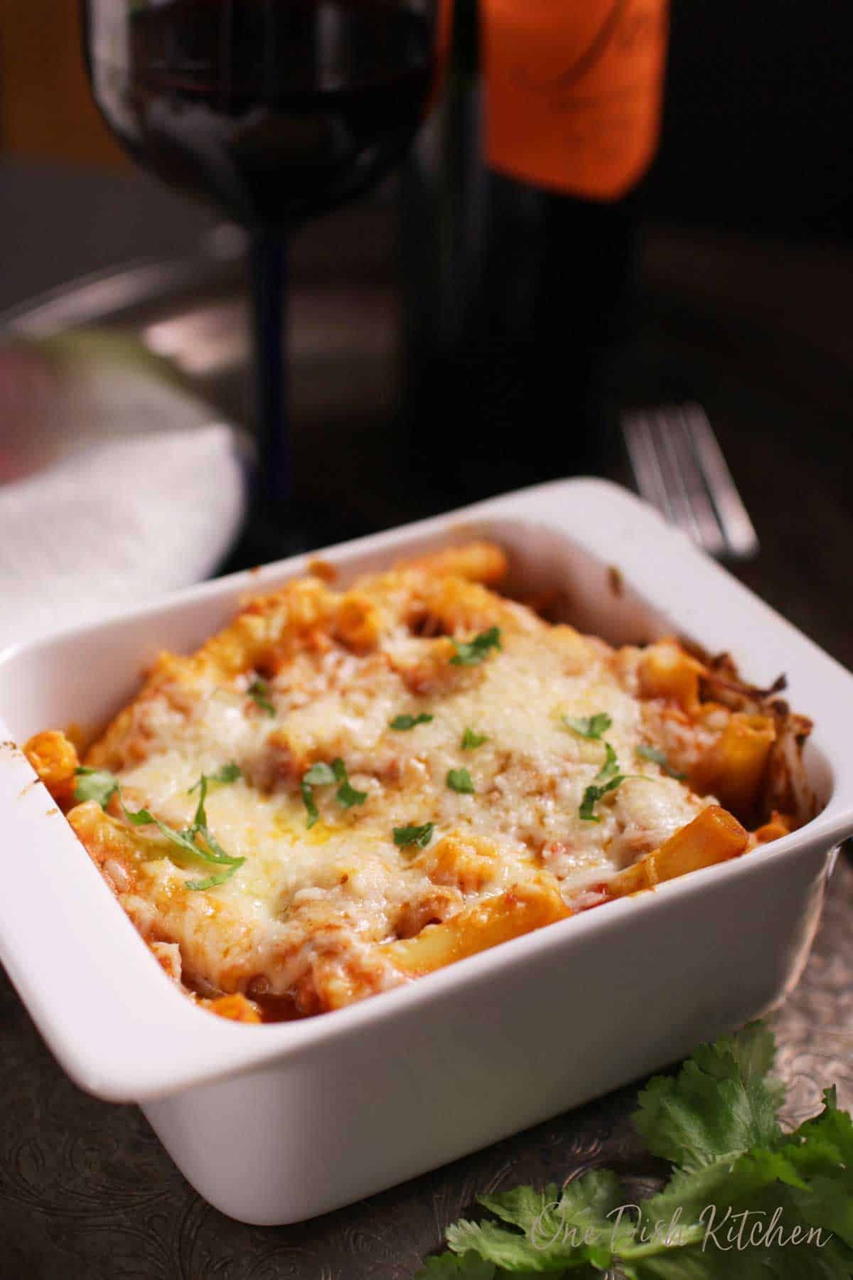 Baked ziti topped with melted mozzarella cheese in a small baking dish garnished with parsley on a metal tray with a glass and a bottle of red wine.