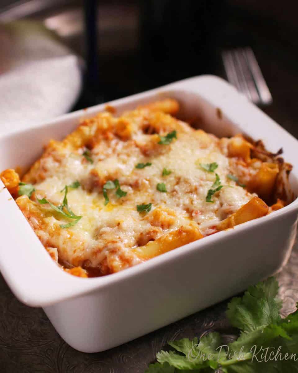 Baked ziti topped with melted mozzarella cheese in a small baking dish garnished with parsley on a metal tray with a glass and a bottle of red wine.