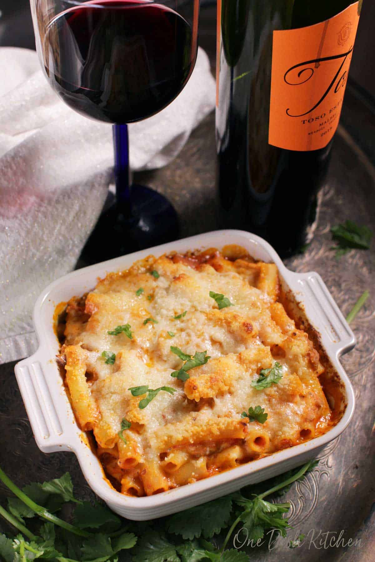 An overhead view of a small baked ziti casserole baked in a small baking dish on a metal tray with a glass and bottle of red wine and parsley. 