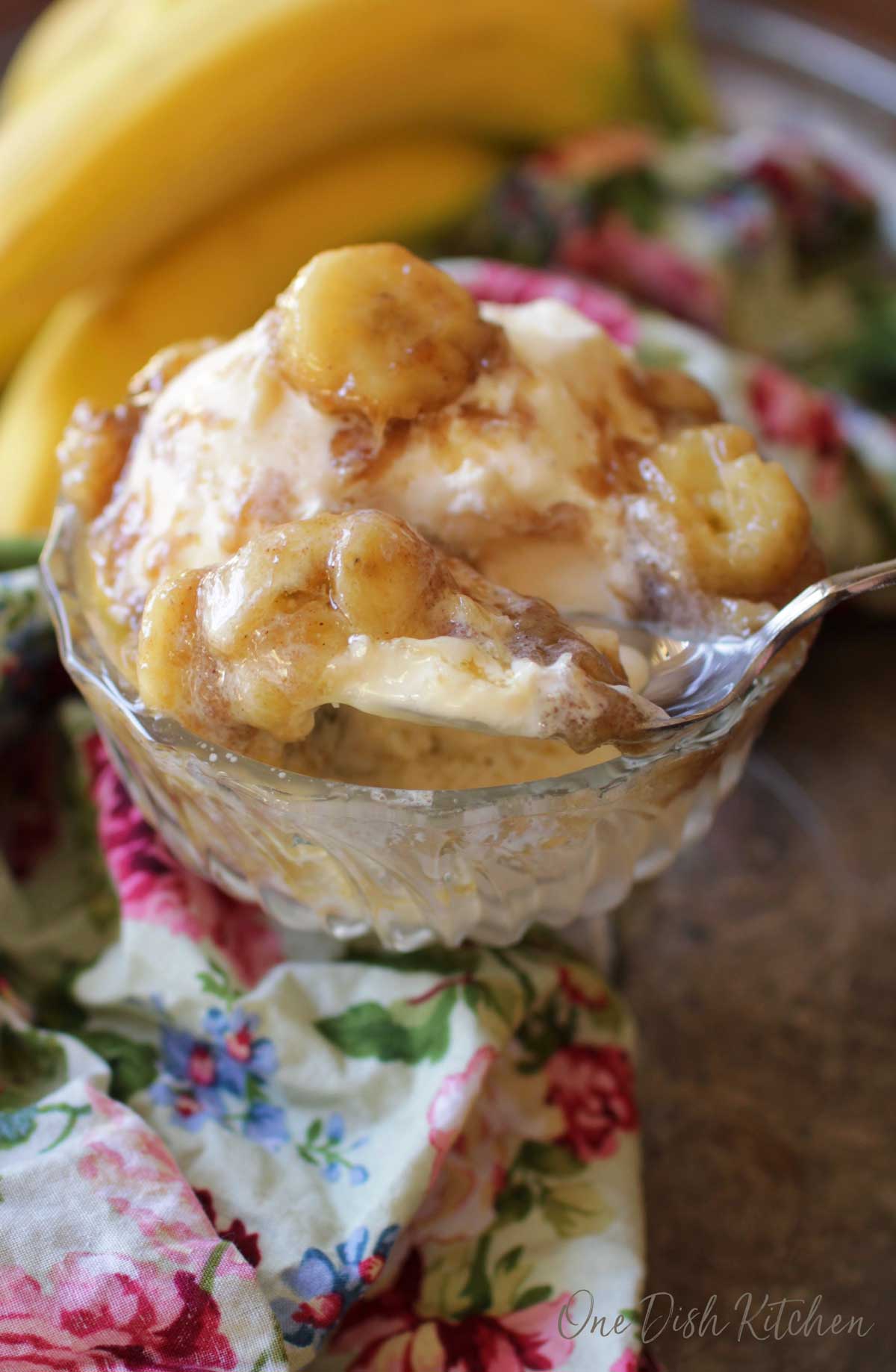 A spoonful of vanilla ice cream that is topped with bananas mixed with brown sugar, butter and vanilla in a dessert glass on a metal tray with a floral cloth napkin and a bunch of bananas.