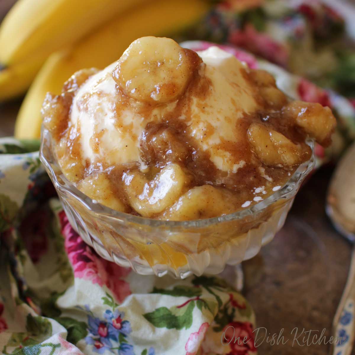 A dessert glass of vanilla ice cream that is topped with bananas mixed with brown sugar, butter and vanilla on a metal tray with a floral cloth napkin and a bunch of bananas. 
