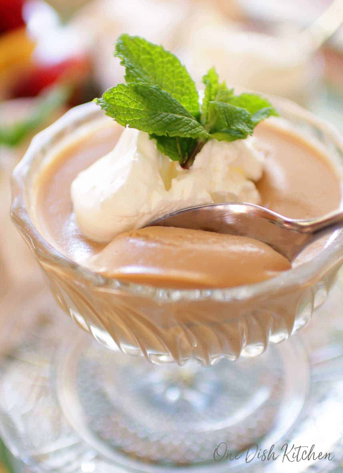 A closeup of a spoonful of butterscotch pudding in a dessert glass topped with whipped cream and mint leaves.