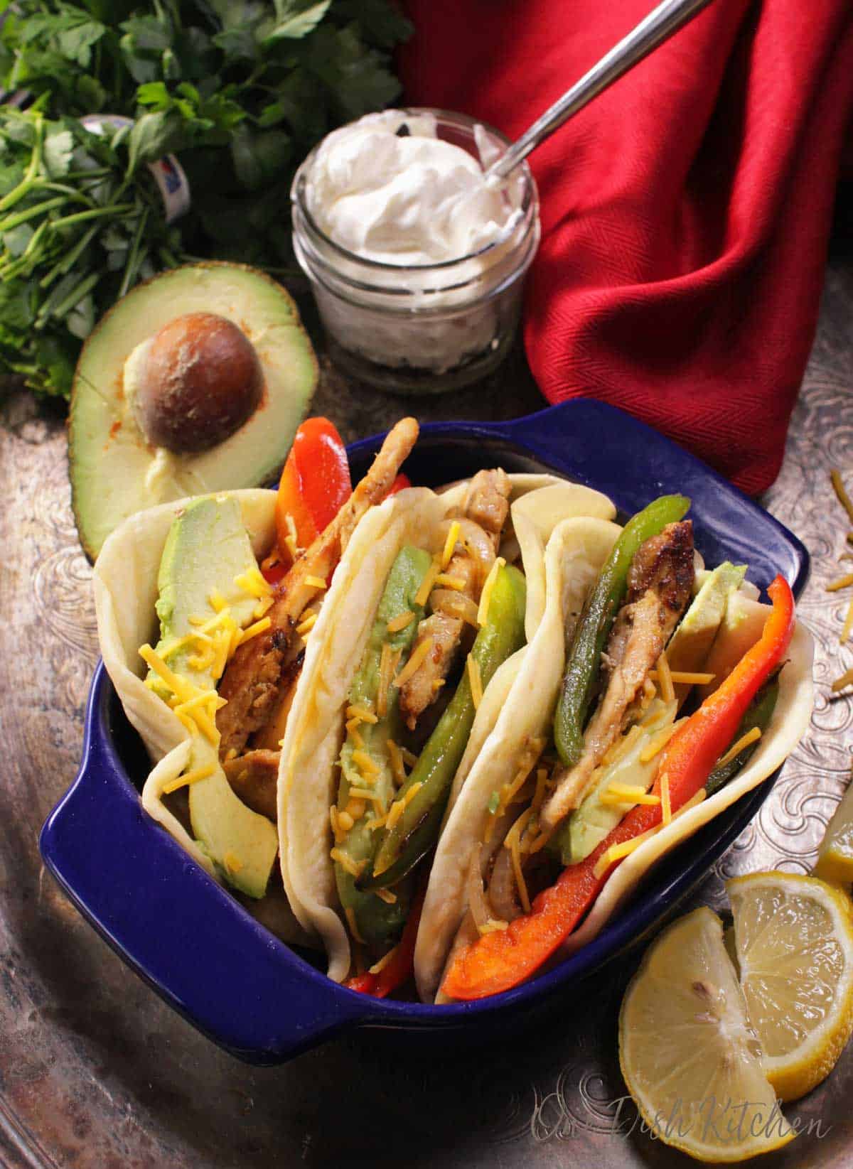 A small baking dish filled with three chicken fajitas filled with avocado slices, red and green bell peppers, and shredded cheese next to a half of an avocado and a small jar of sour cream.