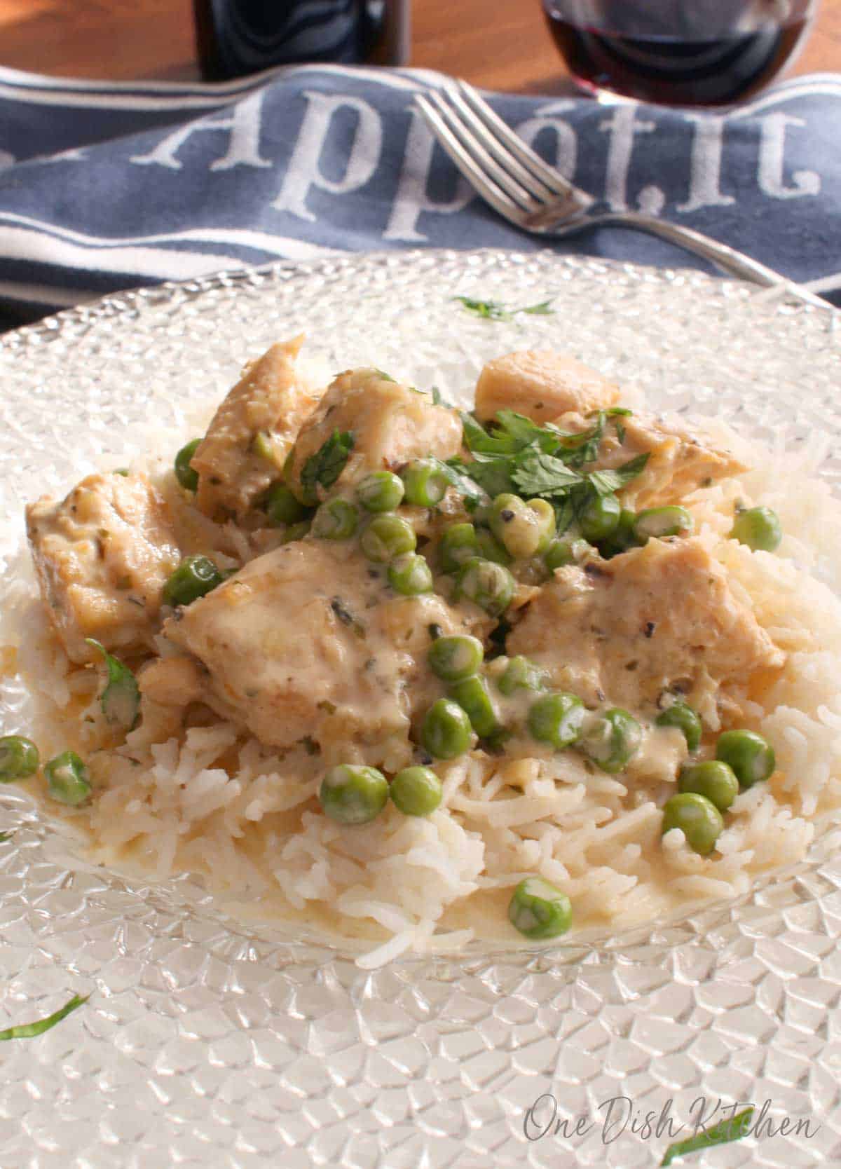 A closeup of a clear glass plate of chicken over white rice with creamy sauce and peas with a blue cloth napkin and a glass of red wine in the background
