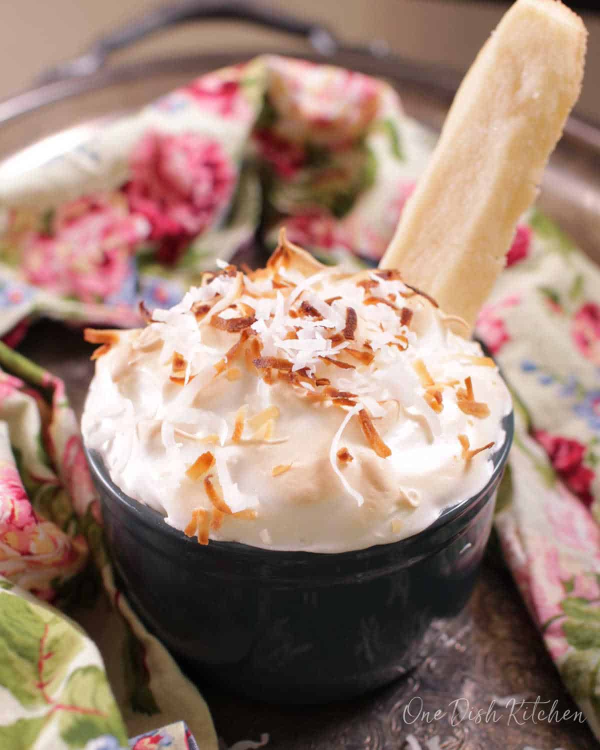 A single serving coconut cream pie in a ramekin topped with coconut shavings next to a floral cloth napkin and a shortbread cookie.
