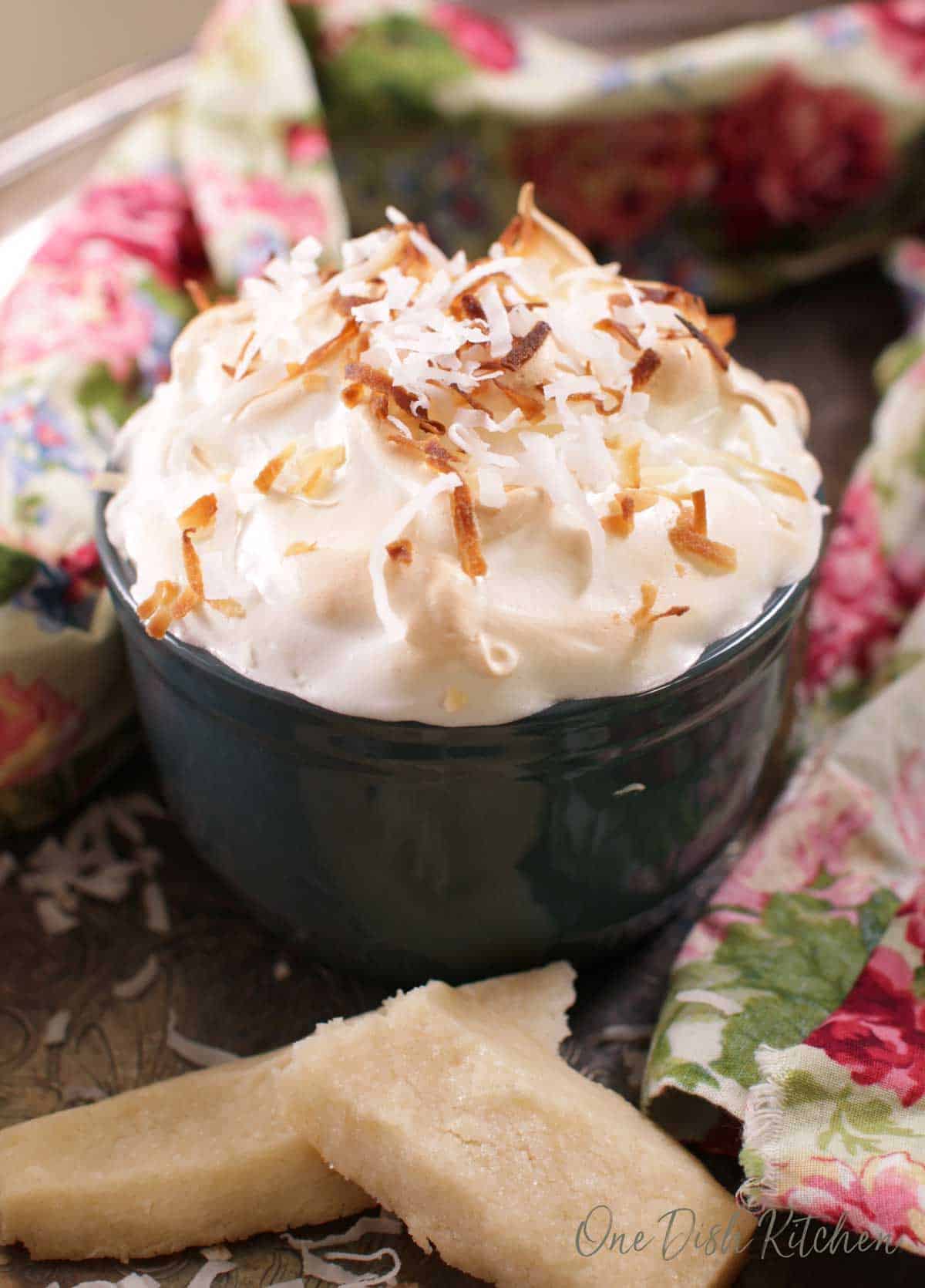 A single serving coconut cream pie in a ramekin topped with coconut shavings next to a floral cloth napkin and a shortbread cookie.