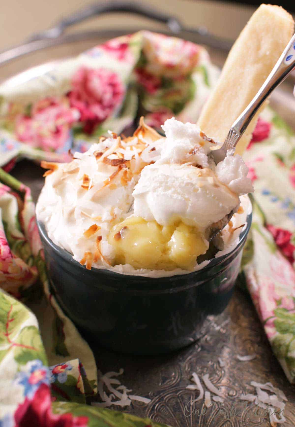 A spoonful of single serving coconut cream pie in a ramekin topped with coconut shavings next to a floral cloth napkin.  
