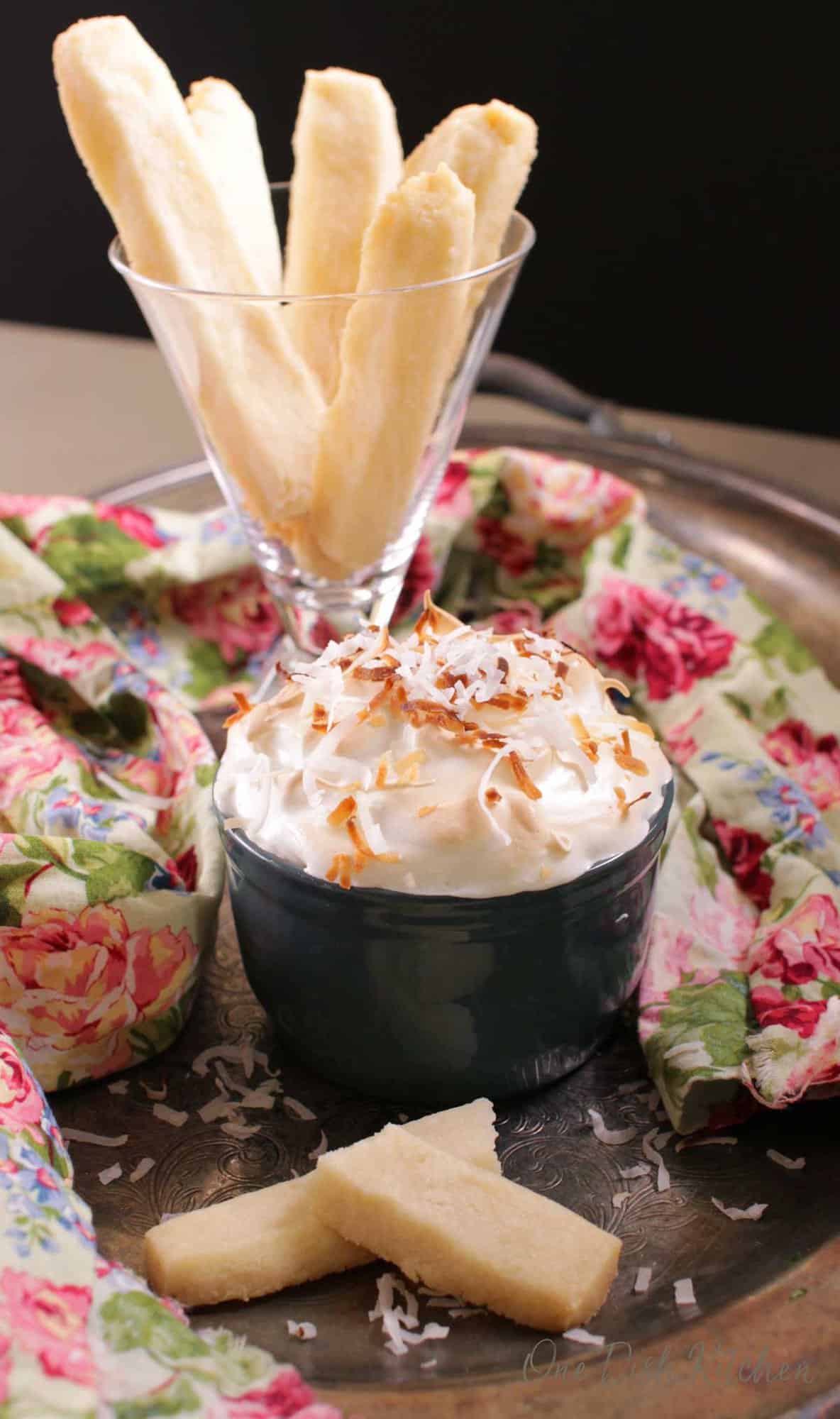 A single serving coconut cream pie in a ramekin topped with coconut shavings next to a floral cloth napkin and a shortbread cookie with a dessert glass of shortbread cookies in the background. 