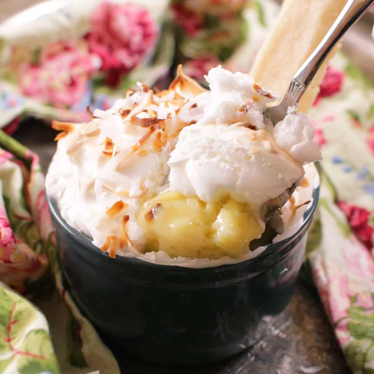 A single serving coconut cream pie in a ramekin topped with coconut shavings next to a floral cloth napkin and a shortbread cookie with a dessert glass of shortbread cookies in the background. 